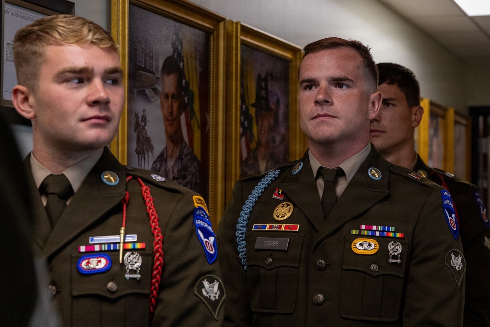 Three military personnel in U.S. Army dress green uniforms are standing in what appears to be a hallway or office corridor. They are wearing various patches, badges, and insignia including Airborne qualifications and unit patches. Framed military portraits hang on the wall behind them.