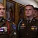 Three military personnel in U.S. Army dress green uniforms are standing in what appears to be a hallway or office corridor. They are wearing various patches, badges, and insignia including Airborne qualifications and unit patches. Framed military portraits hang on the wall behind them.