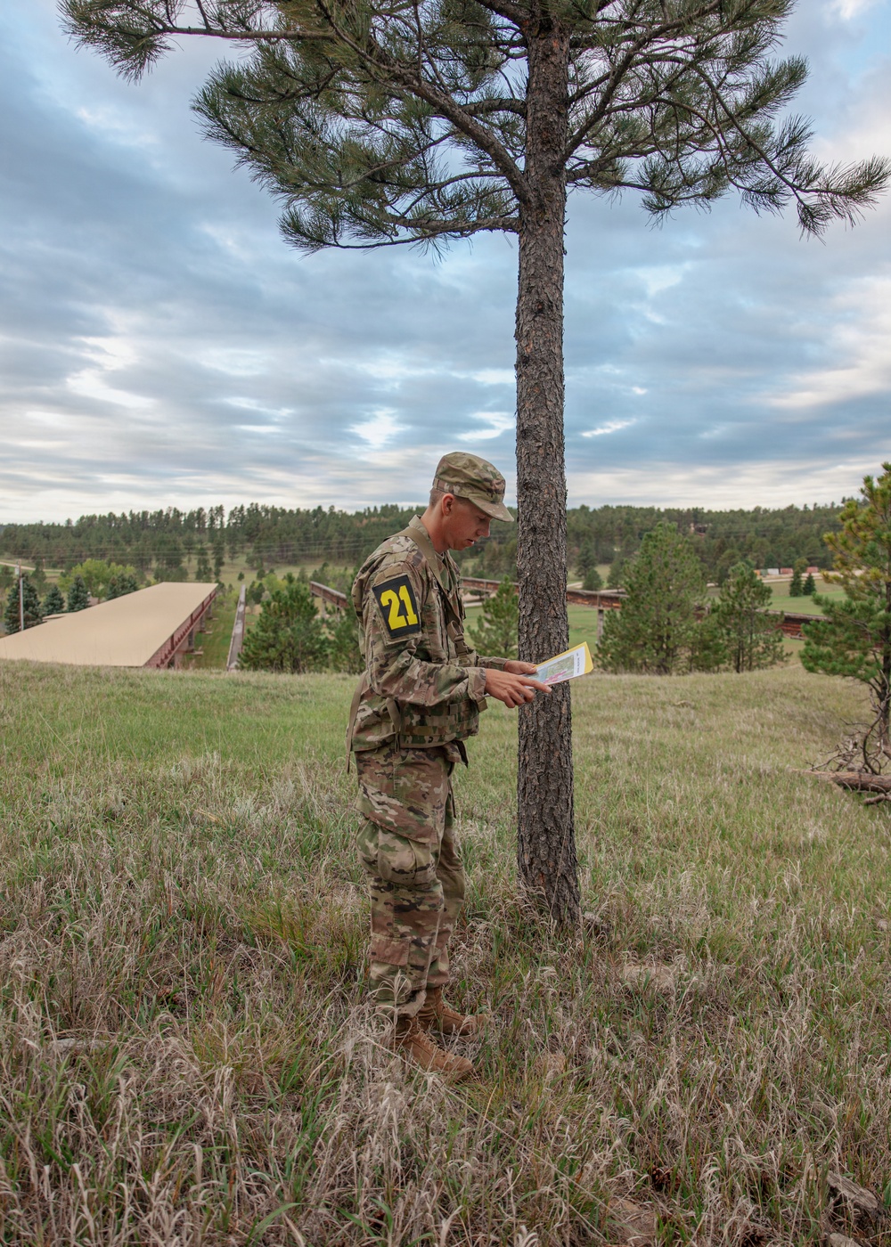 A soldier in US Army OCP (Operational Camouflage Pattern) uniform stands beside a pine tree on an open grassy hillside, consulting what appears to be a map or document. The soldier wears a black patch with the yellow number '21' on the left shoulder, suggesting a unit or competition identifier. The background reveals a military training area with structures, forested terrain, and an overcast sky.