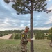 A soldier in US Army OCP (Operational Camouflage Pattern) uniform stands beside a pine tree on an open grassy hillside, consulting what appears to be a map or document. The soldier wears a black patch with the yellow number '21' on the left shoulder, suggesting a unit or competition identifier. The background reveals a military training area with structures, forested terrain, and an overcast sky.