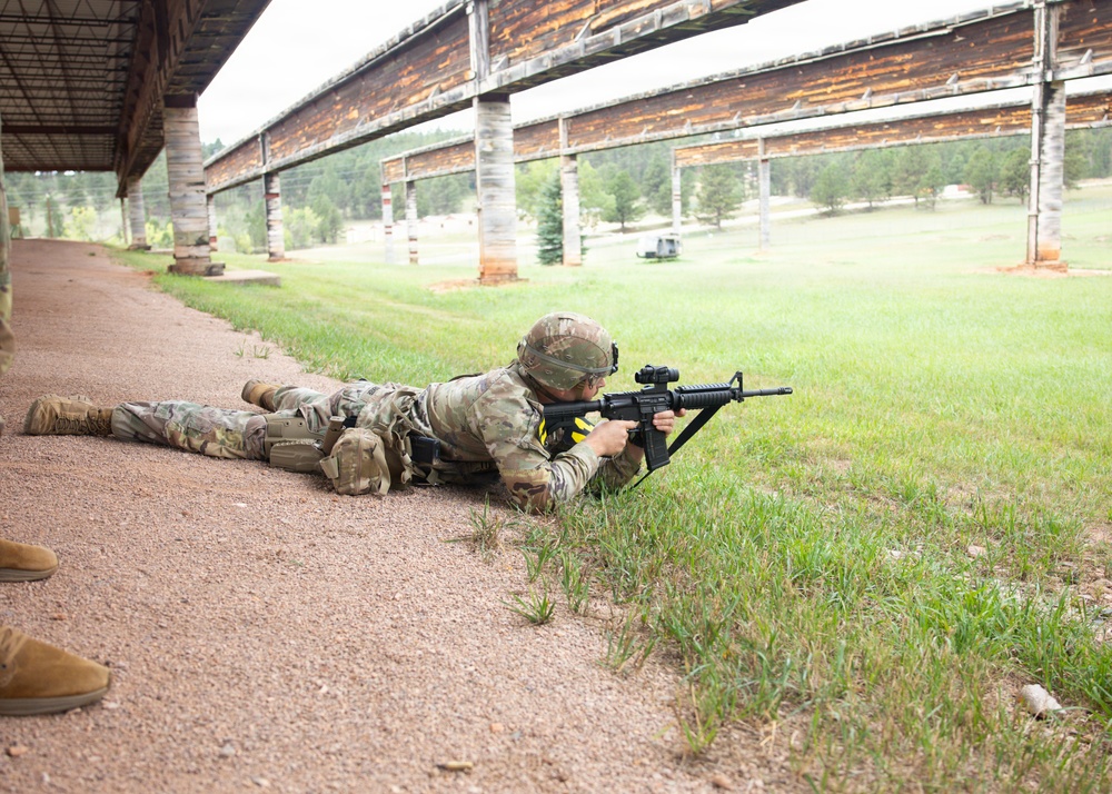 A soldier in full combat gear is in the prone firing position on a military shooting range, aiming a rifle downrange. The setting appears to be an outdoor range with a covered firing line, green grass, and forested hills in the background. The soldier is wearing OCP (Operational Camouflage Pattern) uniform with a helmet and tactical vest.