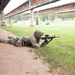 A soldier in full combat gear is in the prone firing position on a military shooting range, aiming a rifle downrange. The setting appears to be an outdoor range with a covered firing line, green grass, and forested hills in the background. The soldier is wearing OCP (Operational Camouflage Pattern) uniform with a helmet and tactical vest.