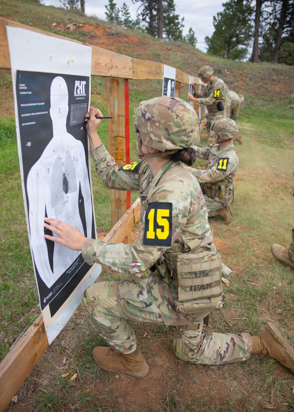 Military personnel in camouflage uniforms are scoring paper shooting targets at an outdoor firing range. At least three individuals wearing numbered yellow identification patches (15, 11, and 14) are kneeling at separate target stands, marking bullet holes on humanoid silhouette targets. The setting appears to be a competitive marksmanship event or qualification exercise in a wooded, hilly environment.