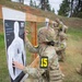 Military personnel in camouflage uniforms are scoring paper shooting targets at an outdoor firing range. At least three individuals wearing numbered yellow identification patches (15, 11, and 14) are kneeling at separate target stands, marking bullet holes on humanoid silhouette targets. The setting appears to be a competitive marksmanship event or qualification exercise in a wooded, hilly environment.