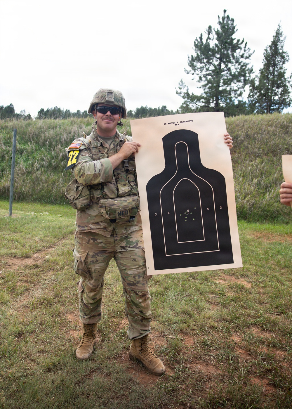 A U.S. Army soldier in full combat gear (OCP/Multicam uniform) holds up a paper shooting target at what appears to be an outdoor firing range. The target is a standard '25 Meter E Silhouette M9' pistol qualification target showing a tight grouping of bullet holes in the center scoring zone. The soldier is wearing the number '22' bib, suggesting this is a competition or qualification event.