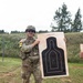A U.S. Army soldier in full combat gear (OCP/Multicam uniform) holds up a paper shooting target at what appears to be an outdoor firing range. The target is a standard '25 Meter E Silhouette M9' pistol qualification target showing a tight grouping of bullet holes in the center scoring zone. The soldier is wearing the number '22' bib, suggesting this is a competition or qualification event.