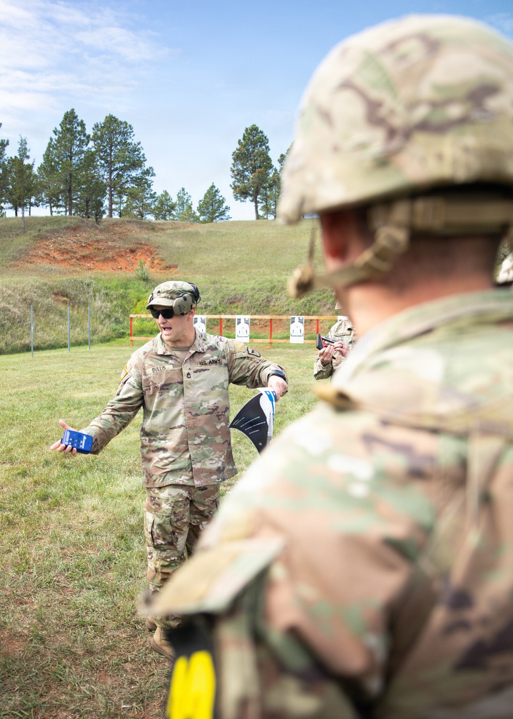 A U.S. Army soldier, identified by name tape as 'BULLIS,' appears to be instructing or briefing other soldiers at an outdoor firing range. He is holding what appears to be a small booklet or card in one hand and a target in the other, gesturing while speaking. Silhouette targets are visible on a berm in the background.