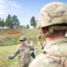 A U.S. Army soldier, identified by name tape as 'BULLIS,' appears to be instructing or briefing other soldiers at an outdoor firing range. He is holding what appears to be a small booklet or card in one hand and a target in the other, gesturing while speaking. Silhouette targets are visible on a berm in the background.
