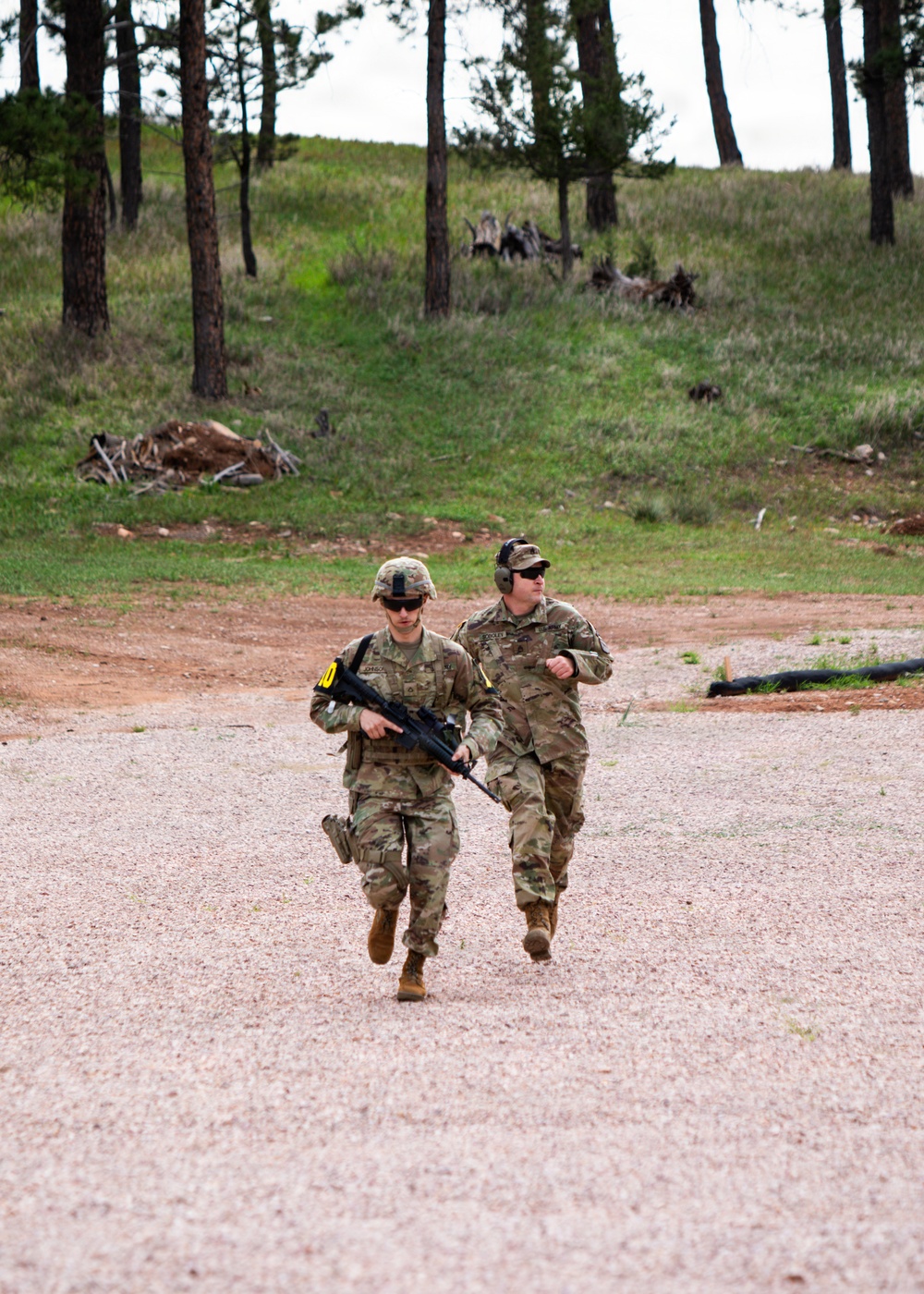 Two military personnel in US Army MultiCam/OCP camouflage uniforms are running along a gravel path in a wooded training area. The lead soldier is carrying what appears to be an M4/M16-style rifle and wearing a helmet with sunglasses, while the second soldier wears a patrol cap with hearing protection. The setting appears to be a military training range based on the terrain, gravel surface, and visible range infrastructure.