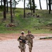 Two military personnel in US Army MultiCam/OCP camouflage uniforms are running along a gravel path in a wooded training area. The lead soldier is carrying what appears to be an M4/M16-style rifle and wearing a helmet with sunglasses, while the second soldier wears a patrol cap with hearing protection. The setting appears to be a military training range based on the terrain, gravel surface, and visible range infrastructure.