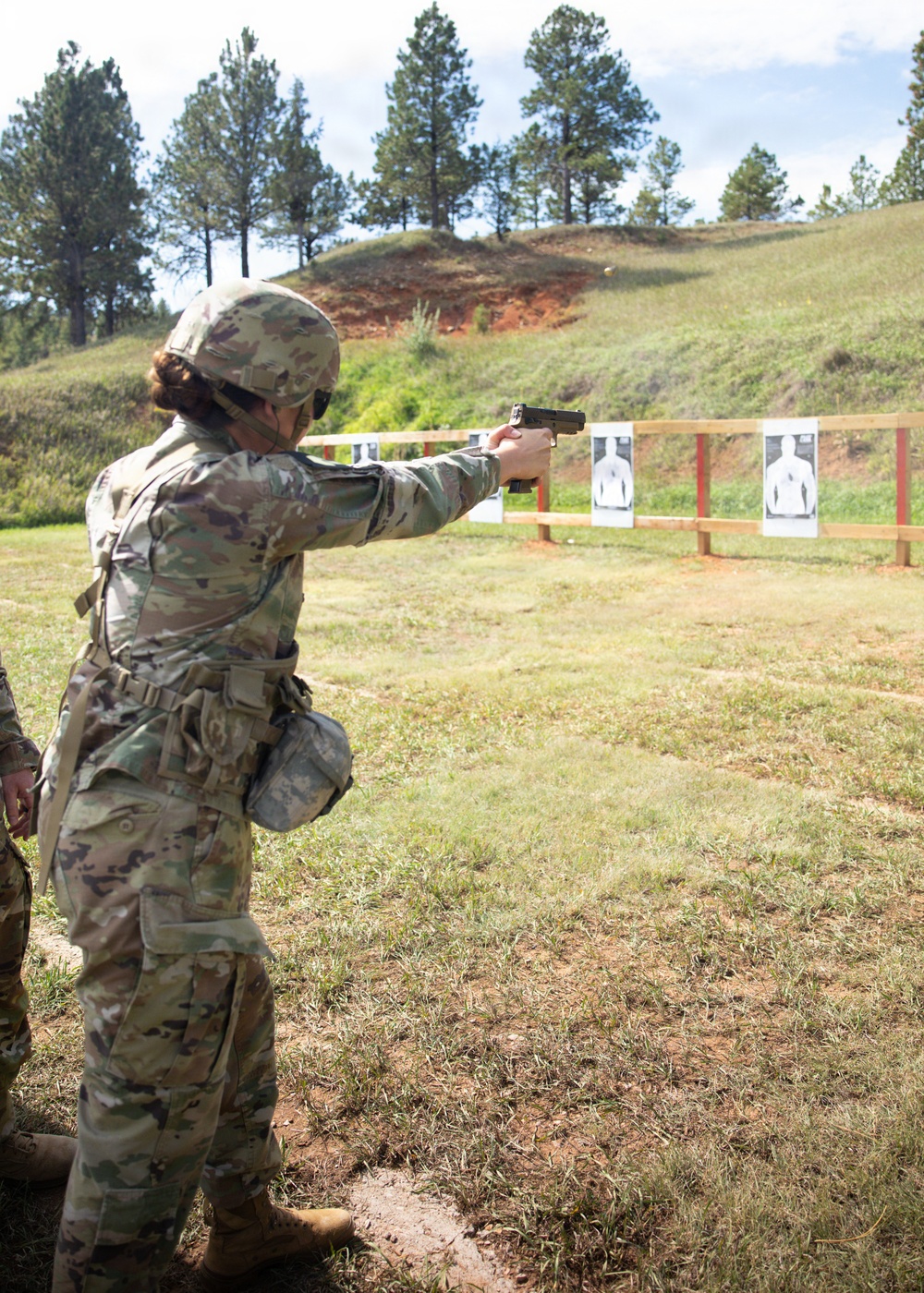 A soldier in full OCP (Operational Camouflage Pattern) uniform and combat helmet is firing a pistol at a military shooting range. The shooter is using a one-handed extended arm stance aimed at silhouette paper targets mounted on a wooden frame. The setting is an outdoor range with a natural earthen berm backstop and pine trees in the background.