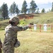 A soldier in full OCP (Operational Camouflage Pattern) uniform and combat helmet is firing a pistol at a military shooting range. The shooter is using a one-handed extended arm stance aimed at silhouette paper targets mounted on a wooden frame. The setting is an outdoor range with a natural earthen berm backstop and pine trees in the background.