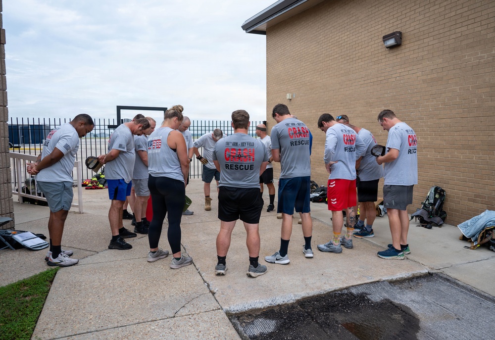 A group of approximately 12-15 individuals wearing matching gray 'Crash Rescue' t-shirts are gathered in a circle with heads bowed, appearing to be in prayer or a moment of reflection. The shirts reference '186th ARW - Key Field' and 'Crash Rescue,' indicating these are Air National Guard crash rescue personnel. The gathering takes place on a concrete pad adjacent to a brick building, likely at Key Field Air National Guard Base in Mississippi.