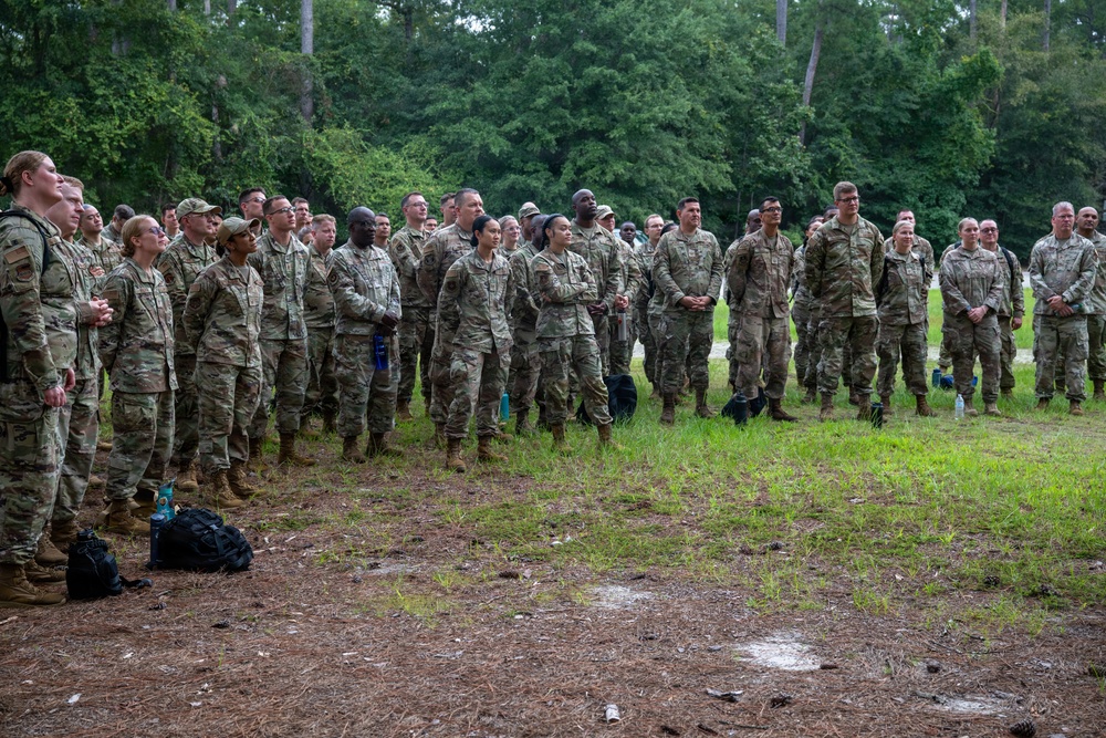 A large group of military personnel in camouflage uniforms are assembled outdoors in a wooded area, standing in a semi-circular formation facing an unseen speaker or focal point. The soldiers appear to be attending a briefing, formation, or ceremony on a grassy and pine needle-covered field. The group is diverse in gender and ethnicity, suggesting a mixed unit gathering.