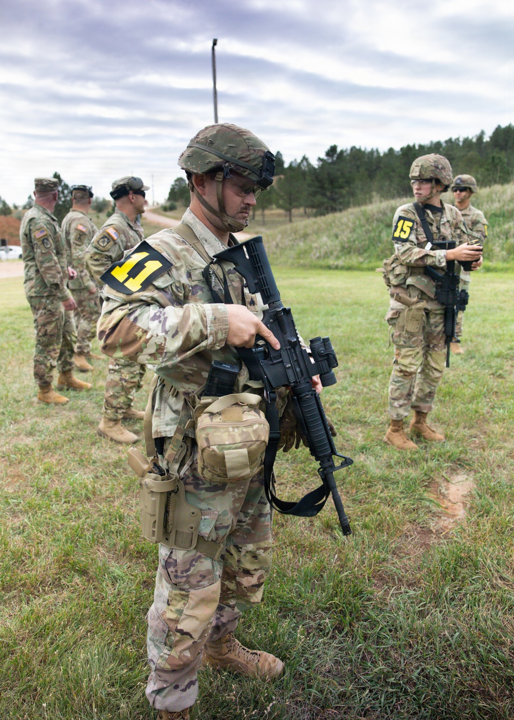 Military personnel in MultiCam/OCP camouflage uniforms are gathered in an outdoor field area, likely during a training exercise. The foreground soldier, numbered '11', is inspecting or demonstrating a rifle equipped with accessories, while other soldiers including one numbered '15' stand nearby. The setting appears to be a military training range or field exercise area.