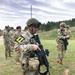Military personnel in MultiCam/OCP camouflage uniforms are gathered in an outdoor field area, likely during a training exercise. The foreground soldier, numbered '11', is inspecting or demonstrating a rifle equipped with accessories, while other soldiers including one numbered '15' stand nearby. The setting appears to be a military training range or field exercise area.