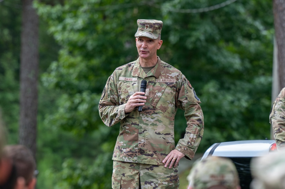 A U.S. Air Force servicemember with the name tape 'DAVIS' is speaking into a handheld microphone, likely addressing a group outdoors. He is wearing OCP (Operational Camouflage Pattern) uniform with a U.S. Air Force tape and a garrison cap. The setting appears to be an outdoor military event or briefing with trees visible in the background.