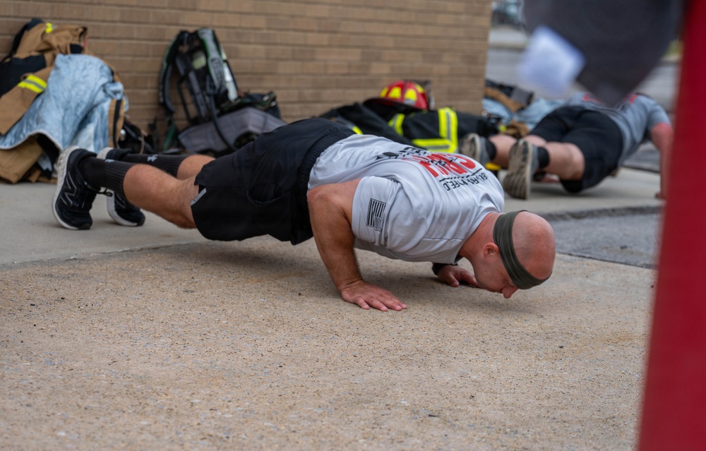 Two individuals are performing push-ups on a concrete surface outdoors, near a brick wall. Firefighting gear, including turnout jackets, helmets, and SCBA equipment, is visible stacked against the wall in the background. The foreground subject is a bald male wearing a white t-shirt, black shorts, and a headband.