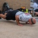 Two individuals are performing push-ups on a concrete surface outdoors, near a brick wall. Firefighting gear, including turnout jackets, helmets, and SCBA equipment, is visible stacked against the wall in the background. The foreground subject is a bald male wearing a white t-shirt, black shorts, and a headband.