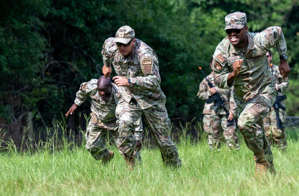 Military personnel in OCP (Operational Camouflage Pattern) uniforms are conducting a field training exercise in a grassy outdoor area. One soldier appears to be assisting or pulling up a fallen comrade, while others run alongside carrying rifles. The scene depicts a dynamic combat or physical training drill.