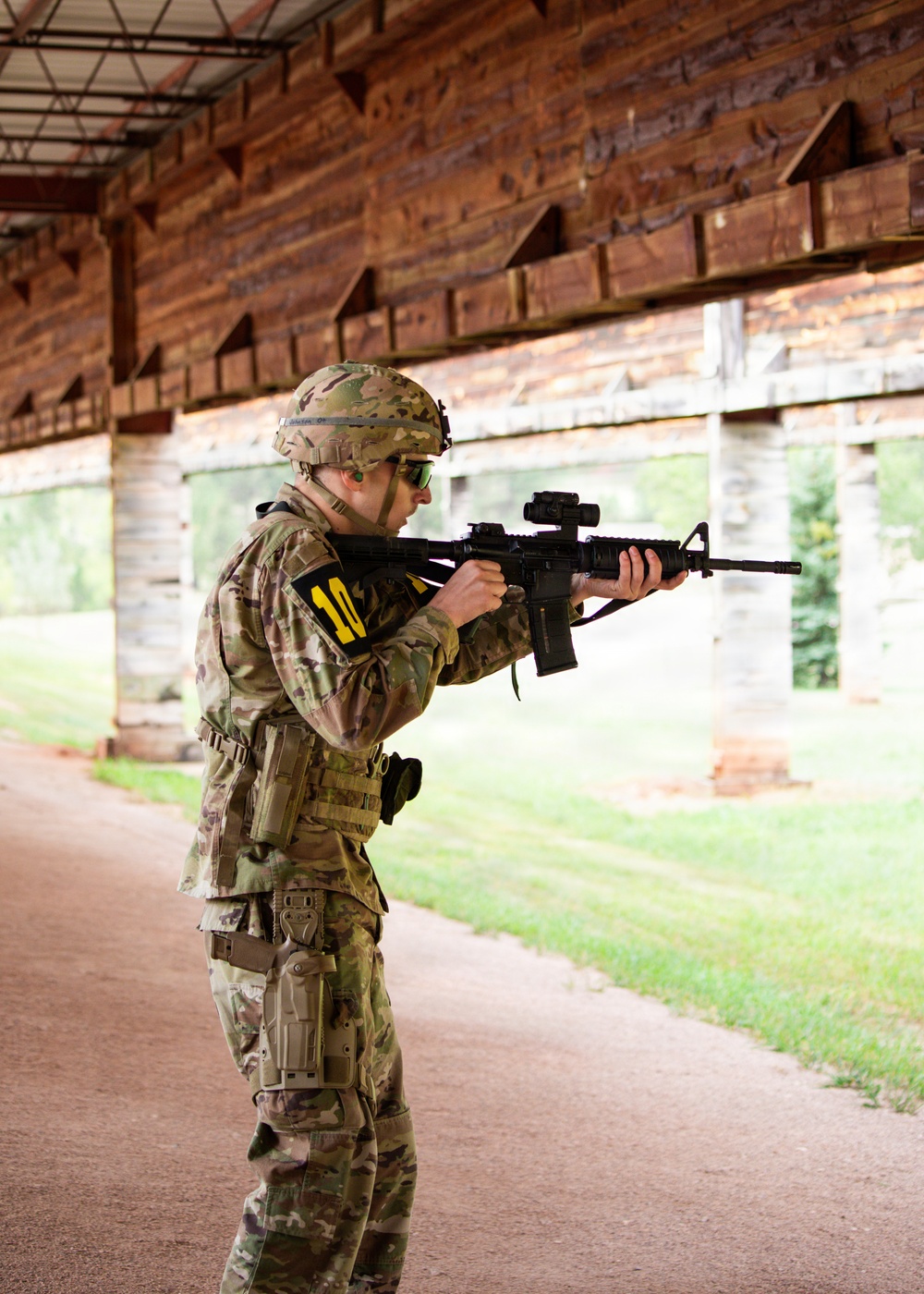 A soldier in full MultiCam/OCP camouflage uniform is in a shooting stance at what appears to be a military firing range. The individual is aiming an M4/M16-style rifle equipped with an optic sight, wearing a combat helmet, protective eyewear, and a thigh holster. A yellow armband with the number '10' is visible on the left arm, suggesting a training exercise or competition.