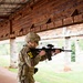 A soldier in full MultiCam/OCP camouflage uniform is in a shooting stance at what appears to be a military firing range. The individual is aiming an M4/M16-style rifle equipped with an optic sight, wearing a combat helmet, protective eyewear, and a thigh holster. A yellow armband with the number '10' is visible on the left arm, suggesting a training exercise or competition.