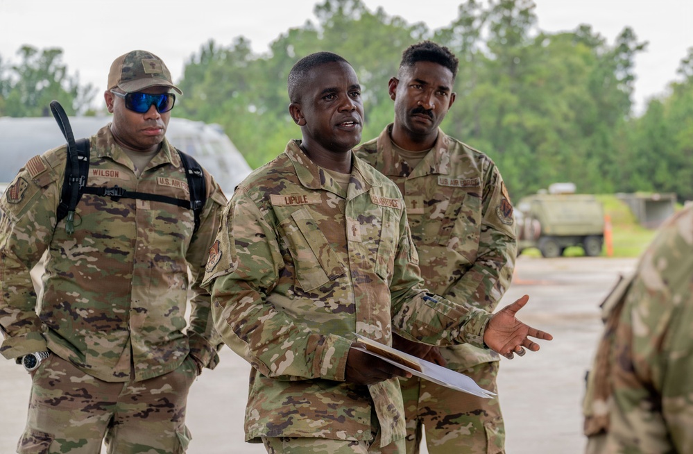 Three U.S. Air Force personnel in OCP (Operational Camouflage Pattern) uniforms are gathered outdoors on what appears to be a military installation or airfield. The central figure, identified by nametape as 'LIPULE,' is speaking and holding documents, suggesting a briefing or coordination activity. A fourth partially visible individual in camouflage is seen in the foreground right.