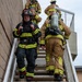 A group of firefighters in full turnout gear are ascending an exterior staircase on the side of a brick building. The lead firefighter wears a black bunker suit and a helmet marked '186' with 'HUNT' visible, while others wear tan/yellow gear with SCBA tanks. This appears to be a training exercise or active emergency response scenario.