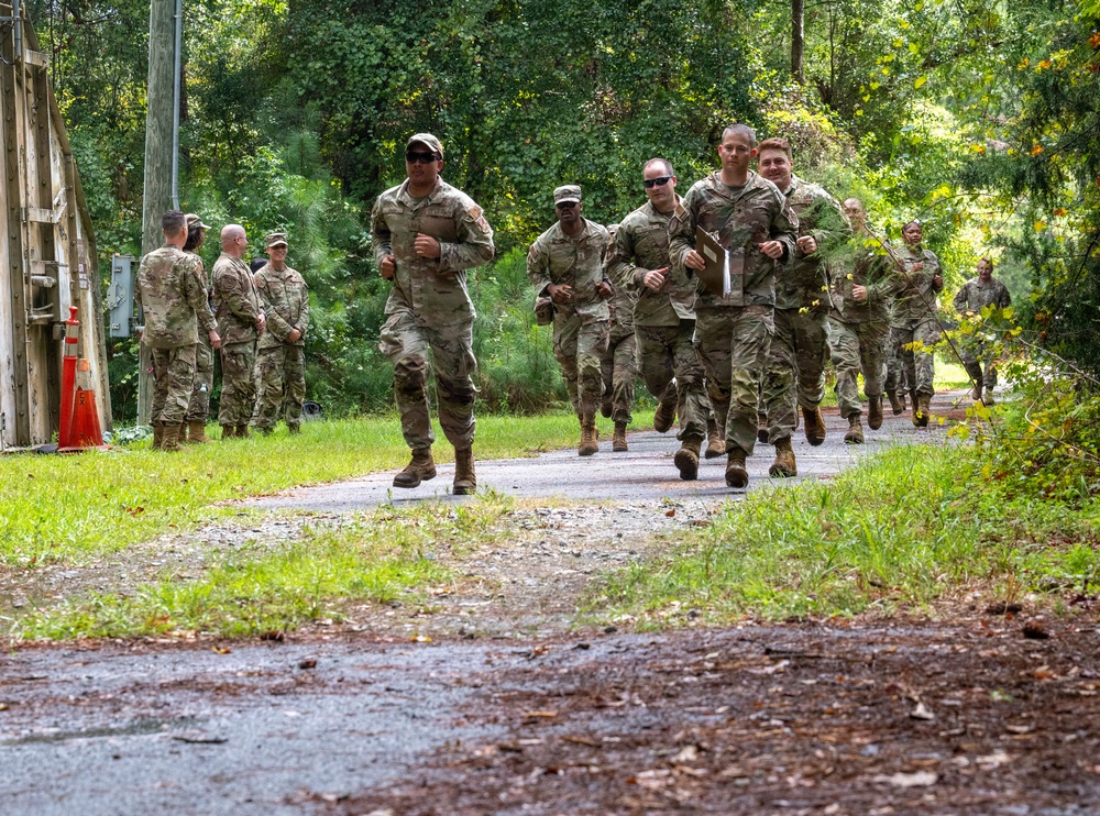 A group of military personnel in U.S. Army OCP (Operational Camouflage Pattern) uniforms are conducting a formation run along a narrow paved path surrounded by trees and grass. Several soldiers are actively running toward the camera while others stand along the left side observing or waiting. The setting appears to be a military installation or training area.