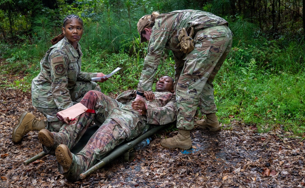Three military personnel in OCP (Operational Camouflage Pattern) uniforms are engaged in what appears to be a field medical training exercise in a wooded outdoor environment. One soldier is lying on a litter/stretcher on the ground simulating a casualty, with visible simulated blood/wounds on the leg, while two others attend to them. One standing soldier holds a clipboard/paperwork, suggesting an evaluator or documentation role, while the other is actively providing simulated medical care.