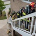 A group of firefighters in full turnout gear are ascending an exterior staircase of a multi-story brick building, appearing to conduct a training exercise or emergency response. The lead firefighter wears a red helmet marked '186' with 'Key Field Crash Rescue Kitchens' visible. Additional firefighters are visible on the ground level below.