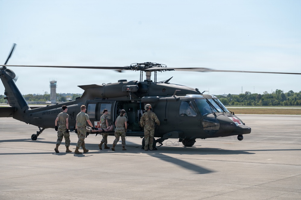 A group of military personnel in camouflage uniforms are loading a patient on a stretcher into a U.S. Army UH-60 Black Hawk helicopter marked with a red cross, indicating a medevac (medical evacuation) mission. The helicopter is on a tarmac at what appears to be a military airfield or joint-use airport, with rotor blades spinning. This appears to be either an actual medical evacuation or a training exercise.