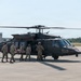 A group of military personnel in camouflage uniforms are loading a patient on a stretcher into a U.S. Army UH-60 Black Hawk helicopter marked with a red cross, indicating a medevac (medical evacuation) mission. The helicopter is on a tarmac at what appears to be a military airfield or joint-use airport, with rotor blades spinning. This appears to be either an actual medical evacuation or a training exercise.