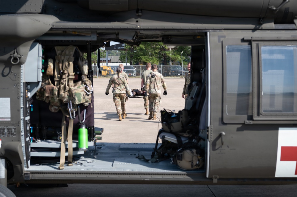 Military personnel in OCP (Operational Camouflage Pattern) uniforms are carrying a patient on a litter/stretcher toward a military medevac helicopter. The image is framed through the open cargo door of what appears to be a UH-60 Black Hawk helicopter configured for medical evacuation, identifiable by the red cross symbol visible on the fuselage. This appears to be a medical evacuation training exercise or operational MEDEVAC procedure on a military installation.