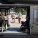 Military personnel in OCP (Operational Camouflage Pattern) uniforms are carrying a patient on a litter/stretcher toward a military medevac helicopter. The image is framed through the open cargo door of what appears to be a UH-60 Black Hawk helicopter configured for medical evacuation, identifiable by the red cross symbol visible on the fuselage. This appears to be a medical evacuation training exercise or operational MEDEVAC procedure on a military installation.