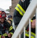A firefighter wearing a red helmet labeled 'Key Field Crash Rescue 186 Kitchens' is holding an ID badge while standing near a staircase railing. He is dressed in full turnout gear with reflective striping and appears to be at an outdoor location near a brick building. Additional firefighters in similar gear are visible in the background.