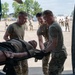 Military personnel in OCP (Operational Camouflage Pattern) uniforms are conducting a medical evacuation drill or training exercise, loading a patient on a litter/stretcher into what appears to be a military aircraft. Four individuals are actively handling the stretcher while a group of uniformed observers watches from the background on a military flight line. The setting appears to be a military airfield or base, likely during a training exercise.