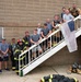 A group of approximately 13-14 firefighters and personnel pose on an exterior staircase of a brick building. Some individuals are in full firefighting gear including turnout coats, helmets, and SCBA equipment, while others are in casual athletic wear. An American flag is draped over the stair railing, and firefighting gear is visible on the ground below.