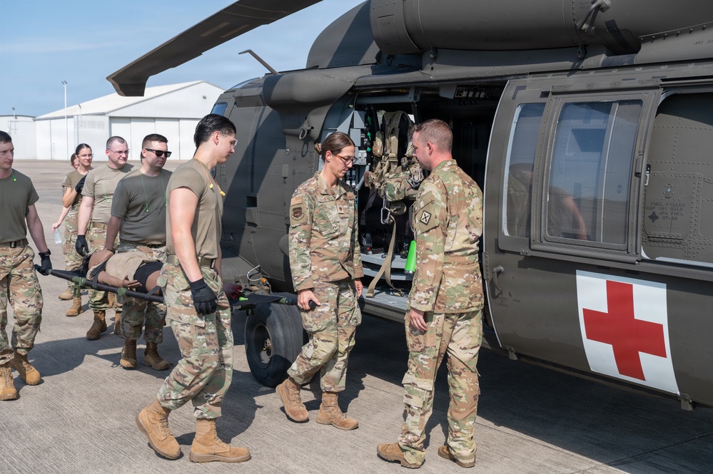 Military personnel are conducting a medical evacuation (MEDEVAC) training exercise near a UH-60 Black Hawk helicopter marked with a red cross, indicating it is configured as a medical evacuation aircraft. Soldiers in OCP (Operational Camouflage Pattern) uniforms are carrying a simulated casualty on a litter/stretcher toward the open side door of the helicopter. A group of additional personnel observes in the background, suggesting this is a training or demonstration scenario.
