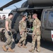 Military personnel are conducting a medical evacuation (MEDEVAC) training exercise near a UH-60 Black Hawk helicopter marked with a red cross, indicating it is configured as a medical evacuation aircraft. Soldiers in OCP (Operational Camouflage Pattern) uniforms are carrying a simulated casualty on a litter/stretcher toward the open side door of the helicopter. A group of additional personnel observes in the background, suggesting this is a training or demonstration scenario.