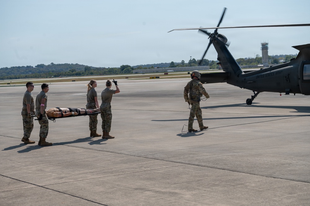 Military personnel in camouflage uniforms are carrying a patient on a litter/stretcher toward a United States Army helicopter on a tarmac. The scene appears to be a medical evacuation (MEDEVAC) exercise or operation, with one soldier signaling toward the aircraft. A control tower is visible in the background, indicating this takes place at a military airfield.