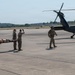 Military personnel in camouflage uniforms are carrying a patient on a litter/stretcher toward a United States Army helicopter on a tarmac. The scene appears to be a medical evacuation (MEDEVAC) exercise or operation, with one soldier signaling toward the aircraft. A control tower is visible in the background, indicating this takes place at a military airfield.
