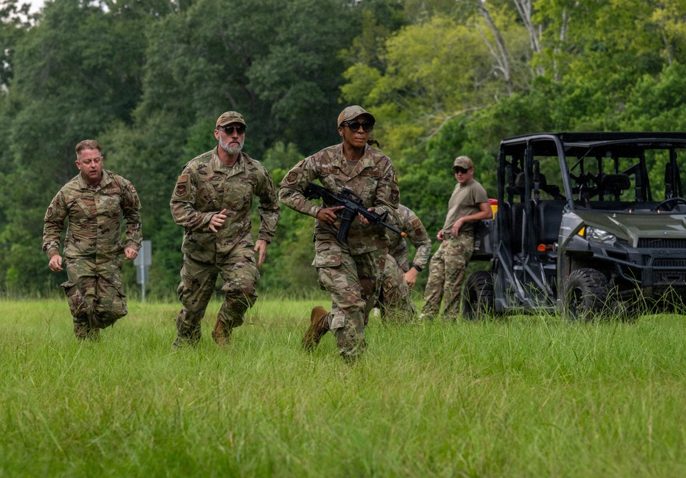 Military personnel in OCP (Operational Camouflage Pattern) uniforms are running across an open grassy field during what appears to be a training exercise. At least one individual is carrying a rifle, and a utility terrain vehicle (UTV) is visible in the background. The setting is a rural or semi-wooded area with dense green tree line in the background.