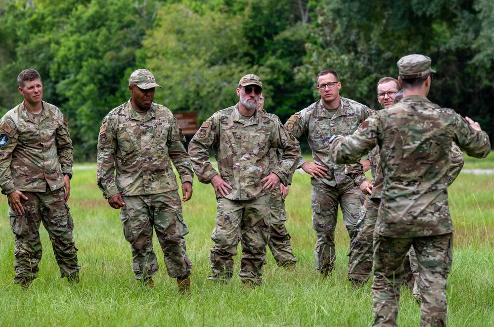 A group of approximately six military personnel in OCP (Operational Camouflage Pattern) uniforms are gathered in an open grassy field with a tree line in the background. One individual facing away from the camera appears to be briefing or instructing the others, who are attentively listening. The setting suggests an outdoor training exercise or field briefing.