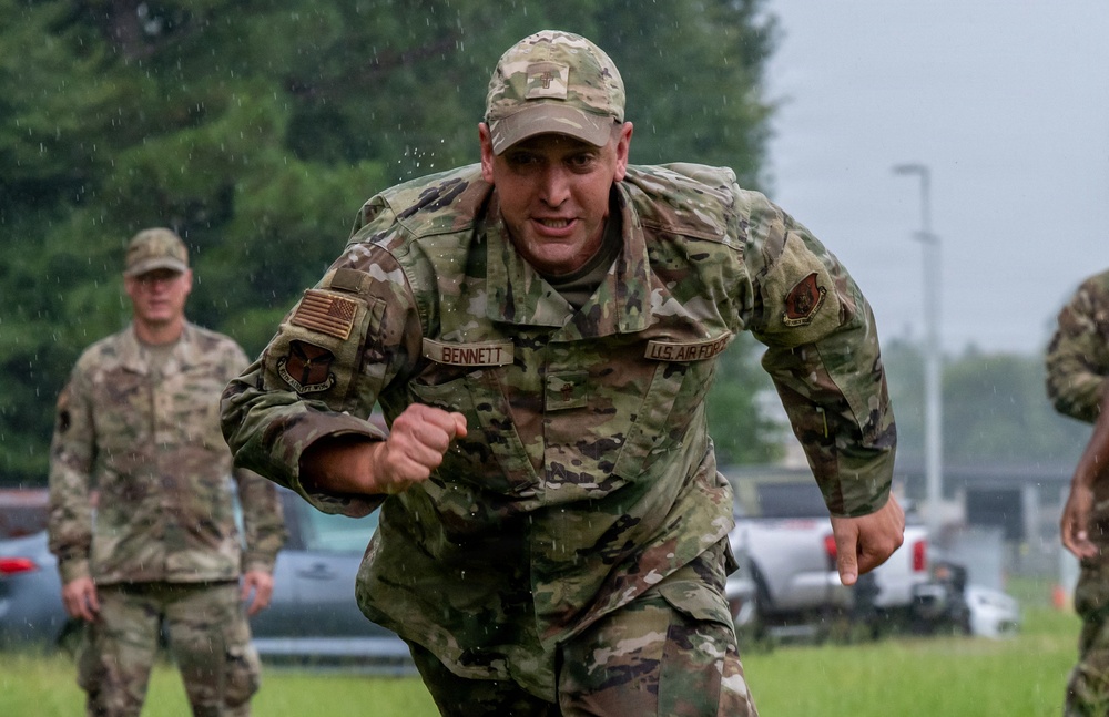 A U.S. Air Force servicemember named Bennett is running intensely in rainy conditions on a grassy field, wearing OCP (Operational Camouflage Pattern) uniform. The individual appears to be participating in a physical fitness event or military training exercise. At least two other uniformed personnel are visible in the background.