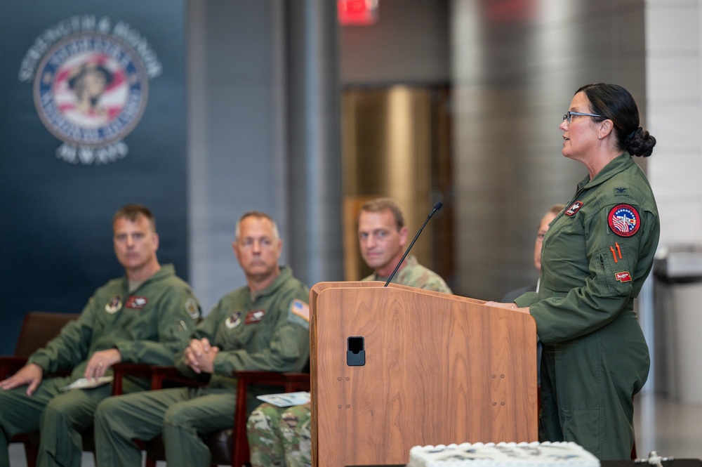 A military officer in olive green flight suit is speaking at a wooden podium during what appears to be an official ceremony or briefing. Three male military personnel in similar flight suits are seated in the background, attentively listening. The setting appears to be an official military facility with an organizational seal/emblem visible on the wall.