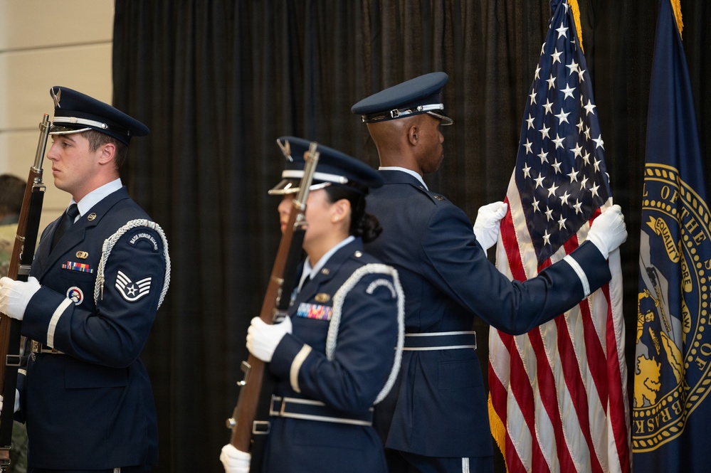 A U.S. Air Force Base Honor Guard color guard team is presenting colors at a formal ceremony. Three uniformed airmen are visible, with one carrying the U.S. flag, one carrying what appears to be a state flag (Nebraska), and one standing at attention with a rifle. The ceremony appears to be taking place indoors against a black curtain backdrop.