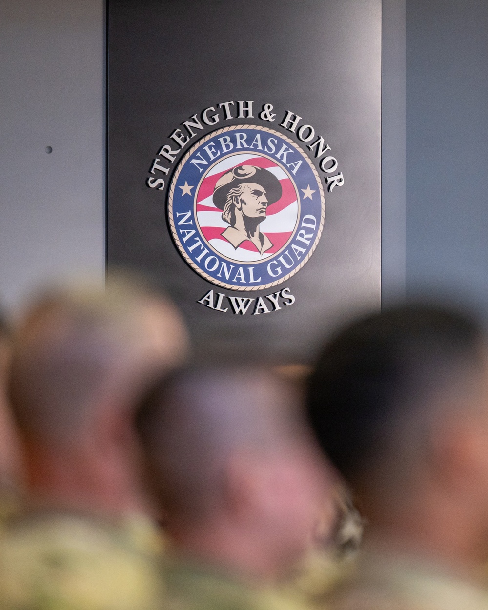 The image shows the official seal of the Nebraska National Guard mounted on a wall, with the motto 'Strength &amp; Honor Always' surrounding it. In the foreground, several blurred figures wearing military camouflage uniforms are visible, suggesting a gathering or meeting is taking place. The seal features a colonial minuteman figure against an American flag background.