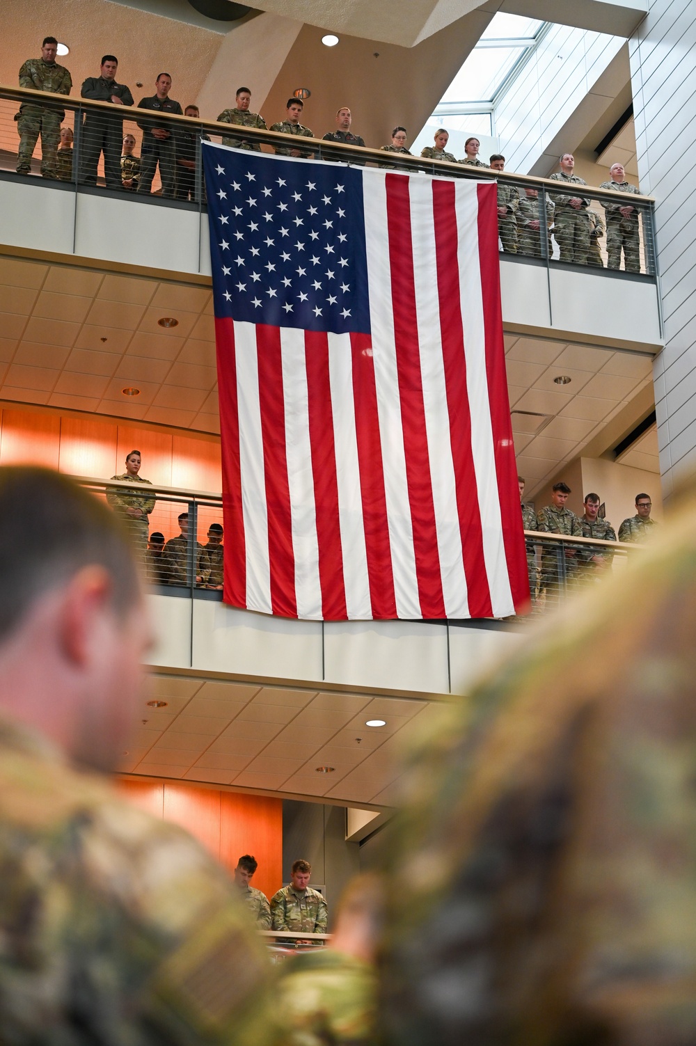 A large American flag is displayed vertically in an indoor atrium of what appears to be a military facility. Numerous uniformed military personnel are gathered on multiple levels/balconies observing a ceremony below. The setting suggests a formal military ceremony or memorial event.