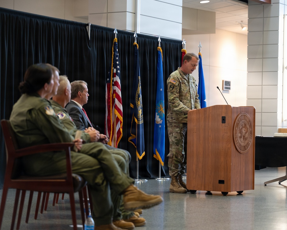 A military ceremony is taking place at what appears to be a Nebraska National Guard facility, as indicated by the seal on the wooden podium. A soldier in Army Combat Uniform (ACU/OCP camouflage) is speaking at the podium while several military personnel and at least one civilian sit in chairs to the left. Multiple flags are displayed in the background, including the U.S. flag, Air National Guard flag, and other military/unit flags.